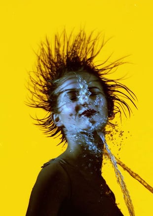 a professionally shot photograph of a model on a yellow background being sprayed in the face with water